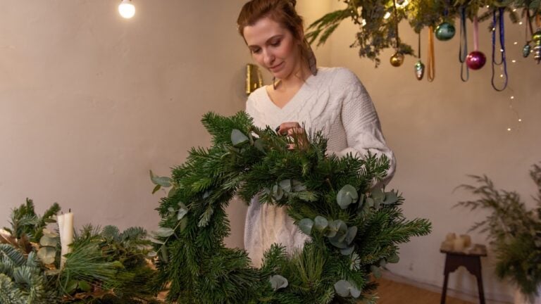 Candid young woman in knitted sweater makes fluffy, green Christmas wreath from natural spruce, pine, eucalyptus branches. Warm, winter atmosphere in interior. Authentic garland of vintage light bulbs