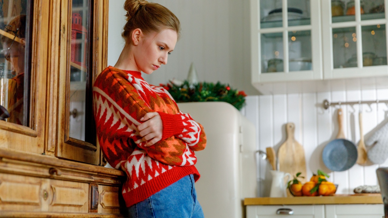 portrait of frowning young blonde woman in red sweater with crossed arms. housewife stands leaning on cupboard in cozy kitchen feeling upset and frustrated