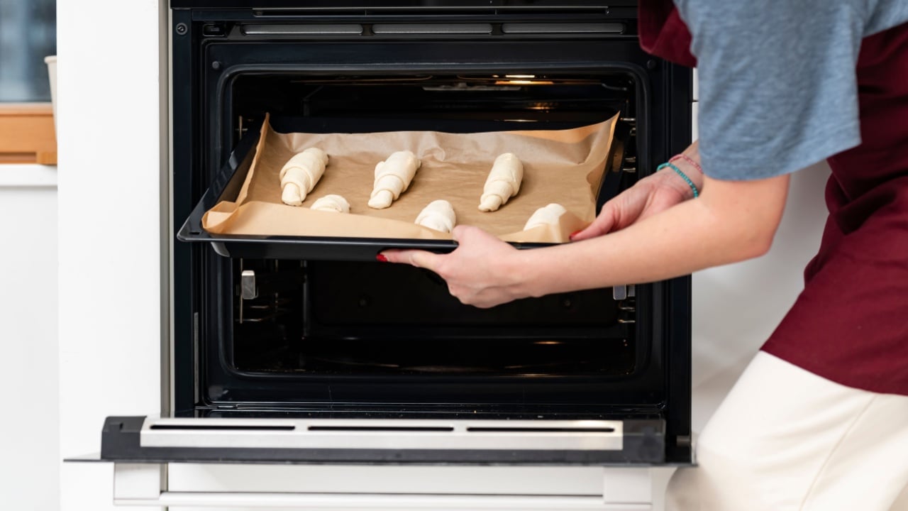 A young housewife in an apron is holding a baking sheet with croissants and is about to put them in the oven