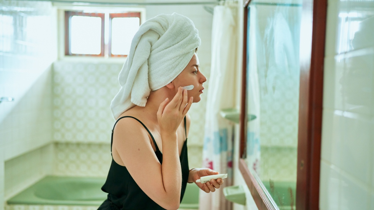 Woman in retro bathroom applies face cream, wrapped in towel. Skin care routine, mirror reflection at home spa. Female enjoys organic skincare product, self-care beauty regimen in vintage interior.