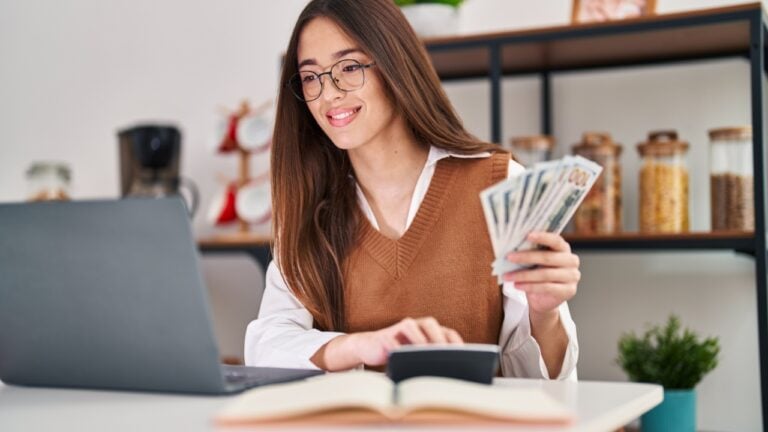Young beautiful hispanic woman using laptop counting dollars at home