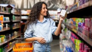 Joyful young woman with curly hair selects products in supermarket, exemplifying healthy lifestyle and shopping habits.