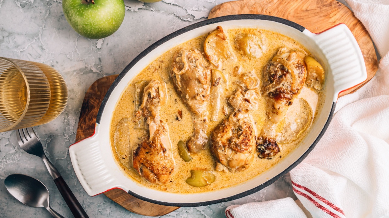 Chicken Normandy served in an enamel-coated cast-iron roaster, apples, glass of cider on a grey marble background. French cuisine. Fall recipe idea