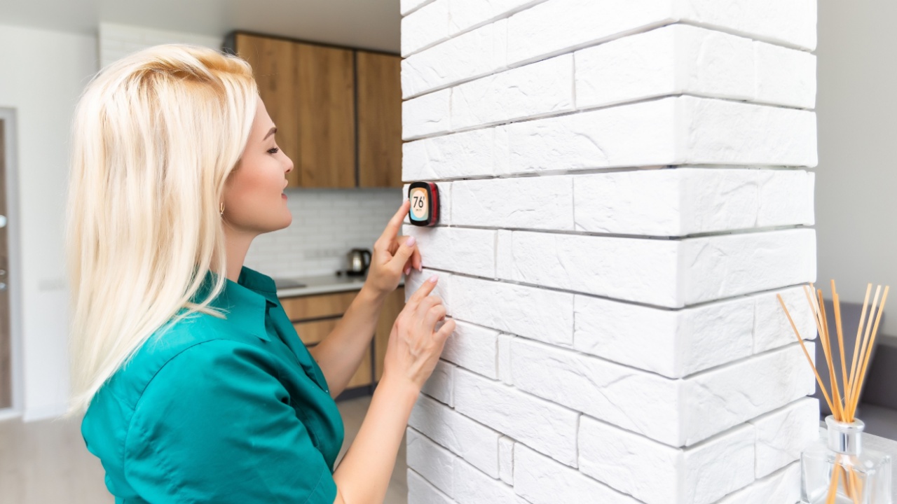 woman regulating heating temperature with a modern wireless thermostat installed on the white wall at home. Smart home heating regulation concept