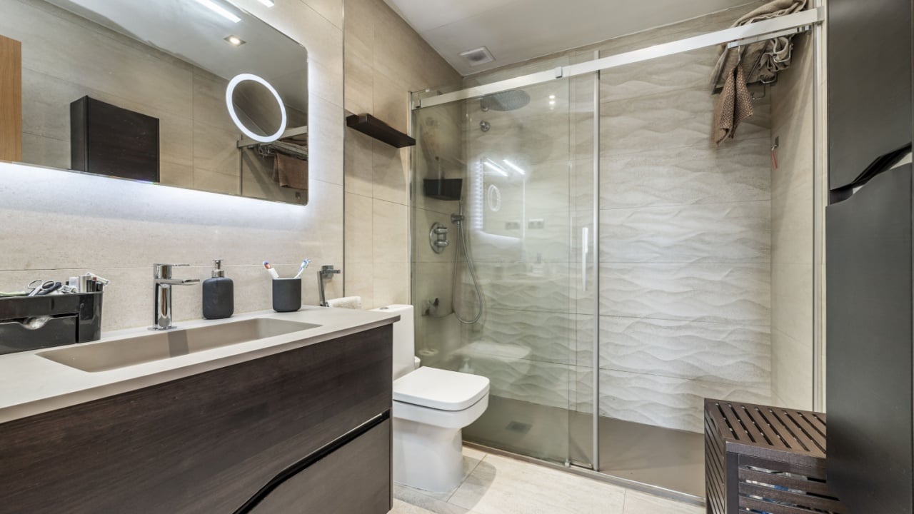 Small bathroom with porcelain toilets, dark cabinet with drawers, mirror with USB lights, square wooden seat and shower cabin with sliding tempered glass screens