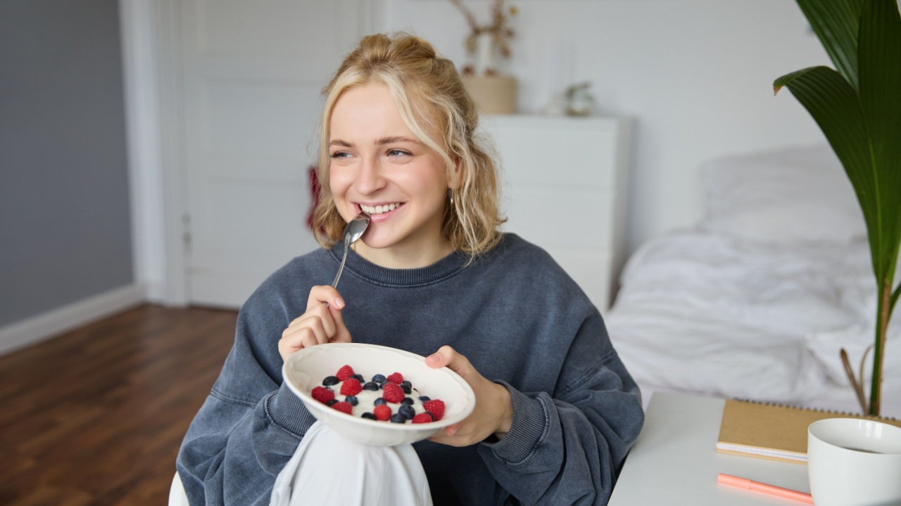 Close up portrait of smiling, cute blond woman, eating healthy lunch in her room, holding bowl with berries, biting spoon, looking happy.