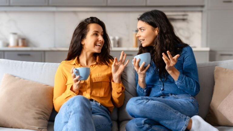 Engaged in a warm conversation, two young pretty women share stories over coffee on a beige couch at home