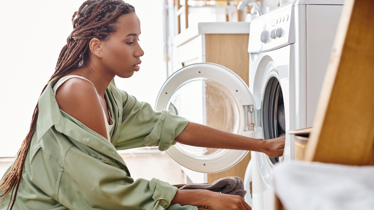 An African American woman with afro braids loads a washer into a dryer while doing laundry in a bathroom.