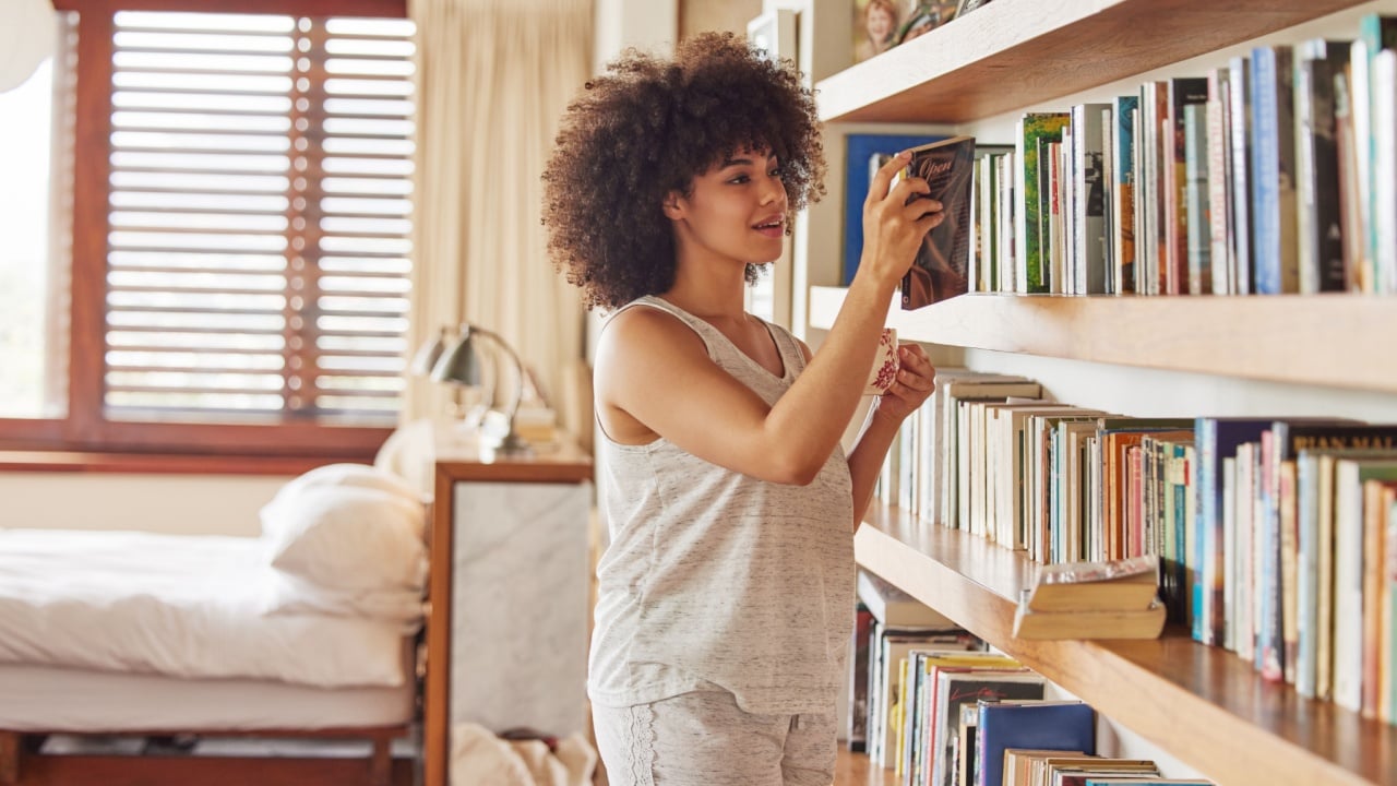 Girl, bookshelf and choice for reading in home, bedroom and morning to relax with thinking on vacation. Woman, person and happy with books, search or decision for story, novel or poetry in apartment