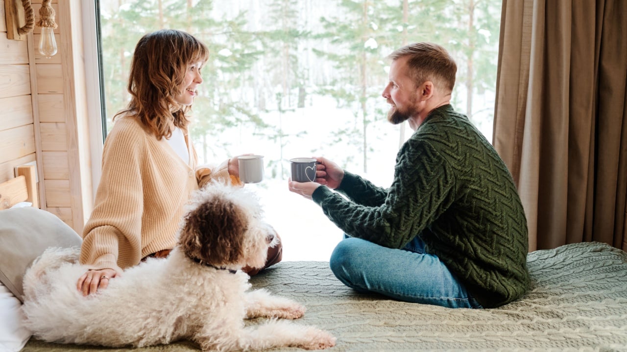 Modern Caucasian man and woman in love wearing casual clothes sitting on bed with their dog on it, drinking tea and talking