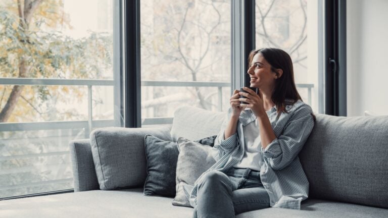 Cheerful dreamy girl holding mug of hot beverage, looking away in deep good thoughts, smiling, dreaming, enjoying aromatic herbal tea, smelling morning coffee, warming hands