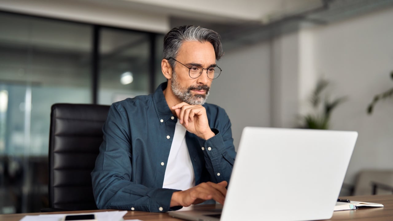 Busy middle aged business man executive wearing shirt and glasses sitting at desk using laptop. Mature serious professional businessman manager working looking at computer technology in office.