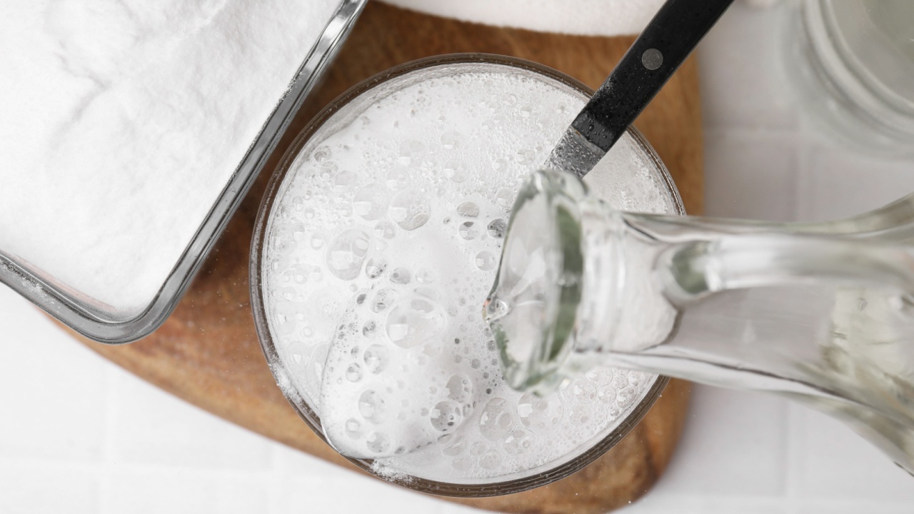 Pouring vinegar into spoon with baking soda over bowl at white tiled table, top view