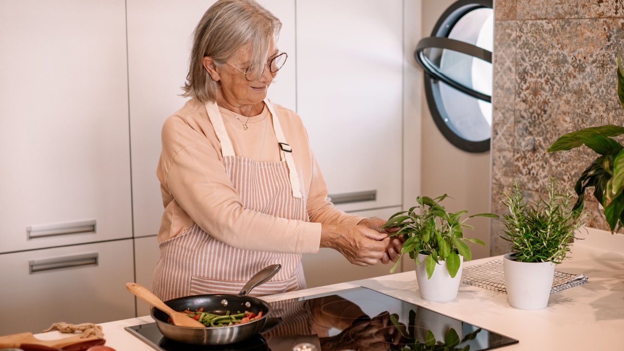 Smiling senior gray haired woman at home cooking vegetables in the pan adds some herbs. People in the kitchen preparing dinner. An elderly woman works in the kitchen