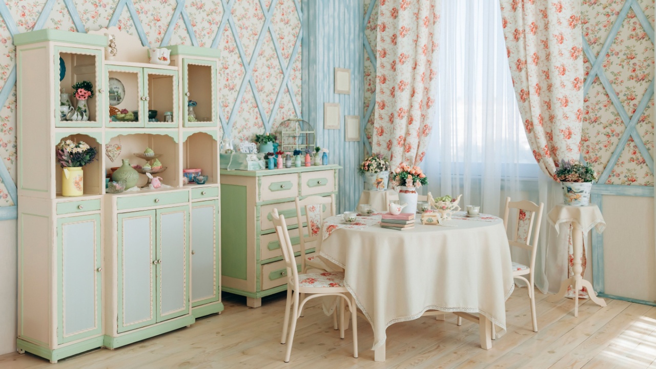 Vintage kitchen with painted pastel green and cream furniture, floral wallpaper, and lace curtains. Cozy dining area with round table and floral decor. Wooden floor enhances rustic charm and warmth