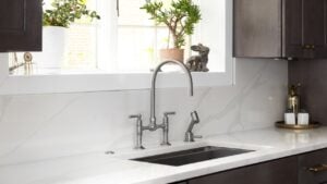 A kitchen faucet detail in a cozy kitchen with a stainless faucet, white marble countertop and backsplash, a bright window, and dark wood cabinets.