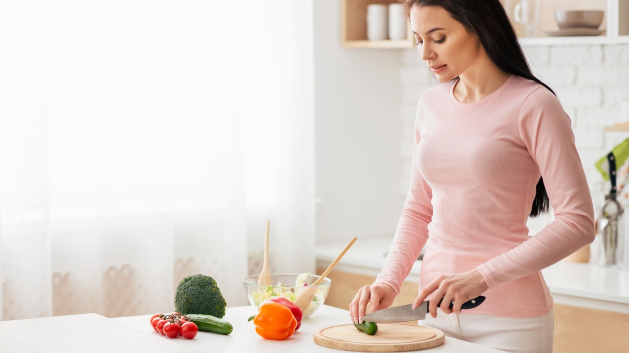 A woman is standing at a kitchen counter, focused on cutting various vegetables on a wooden cutting board. She is using a sharp knife to chop carrots, bell peppers, and onions into small pieces