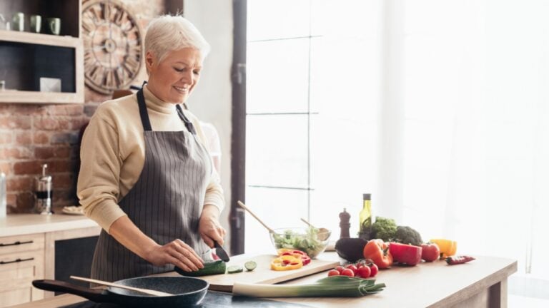 A senior woman with short gray hair is wearing an apron and is cutting vegetables on a cutting board in a modern kitchen. She is smiling as she prepares the vegetables