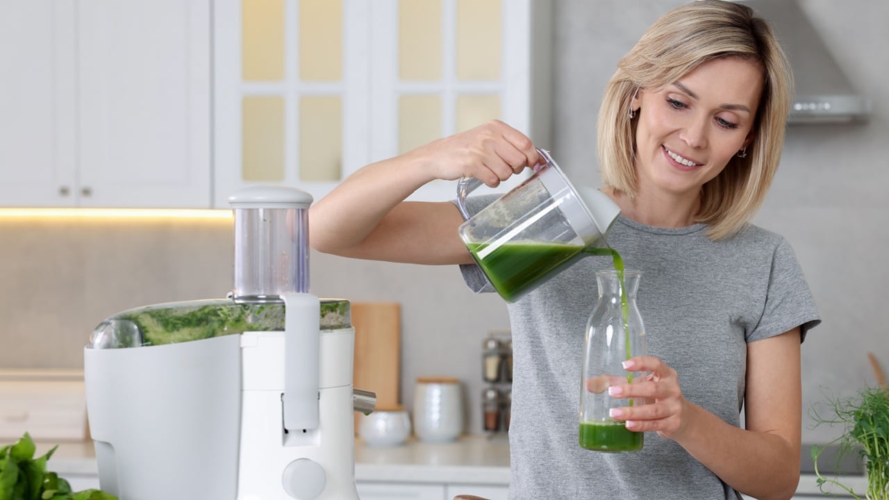 Smiling woman pouring juice into jug in kitchen. Juicer and fresh products on white marble table