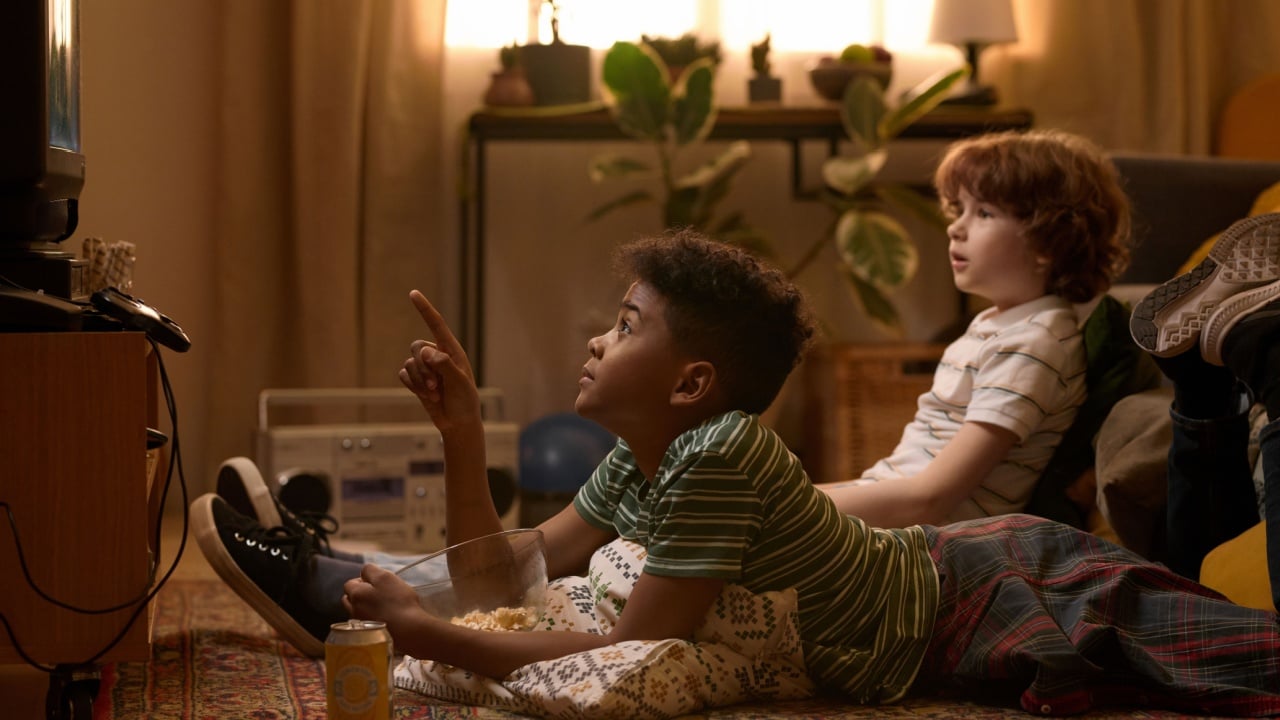 Two multiethnic schoolkids lying on floor, one pointing at TV while watching exciting movie in cozy retro-styled living room with relaxed atmosphere and snacks like popcorn and soda