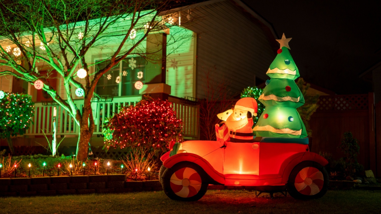 Luminous inflatable figure of Santa Claus in red car with pine tree on the decorated house background