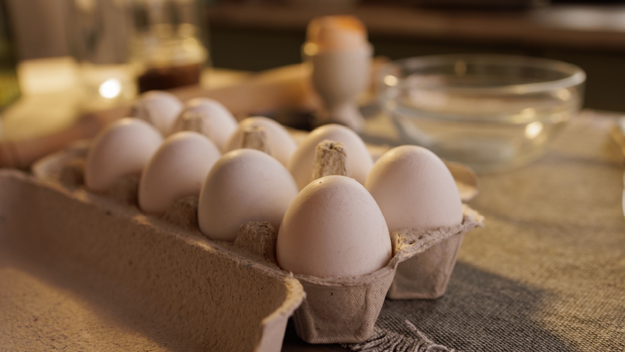 A close-up of a carton of white eggs on a kitchen countertop, with a blurred background showing a person and kitchen utensils