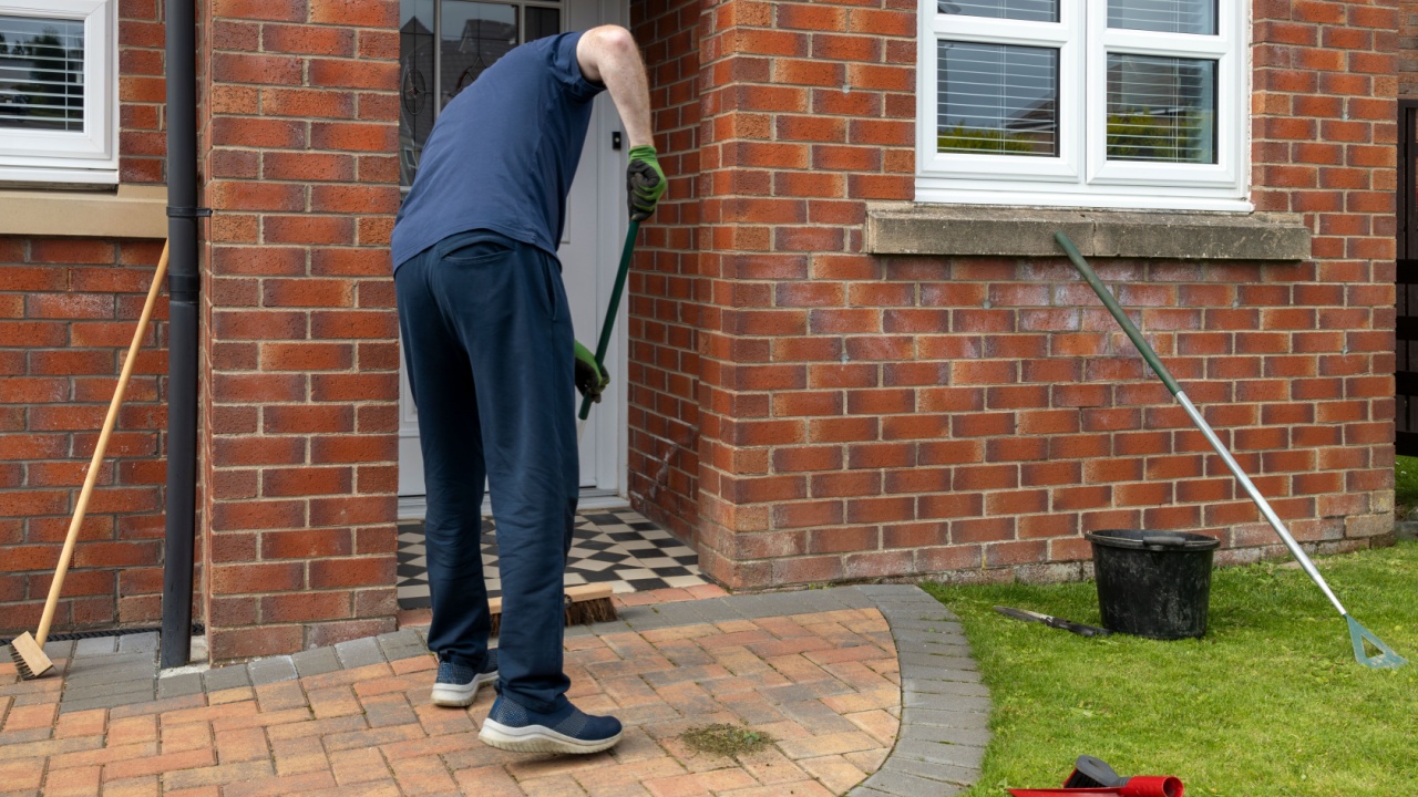 Man sweeping a brick driveway path in front of a house with bucket, broom, and other cleaning tools nearby