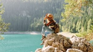 Traveler woman wearing a stylish hat and sweater enjoying a scenic lakeside landscape in the mountains during sunny autumn weather. Eco tourism. Active lifestyle.