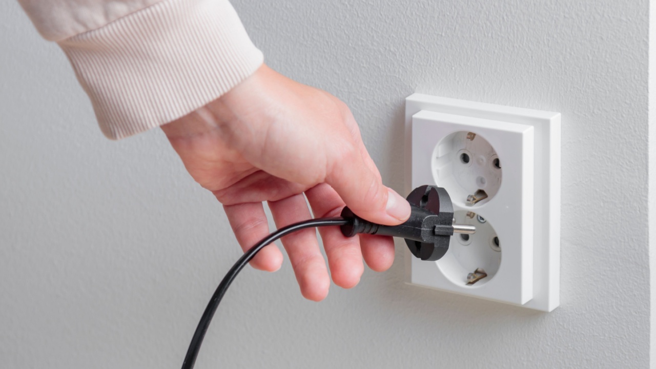 A woman's hand unplugging a power cord from an electrical outlet on a white wall. Careful resource use, the rising cost of electricity and the need for mindful consumption