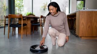 Happy African American woman kneels on the floor, operating a robotic vacuum cleaner in kitchen. The woman uses the vacuum cleaner at home. Housewife using vacuum cleaner. Cleaning