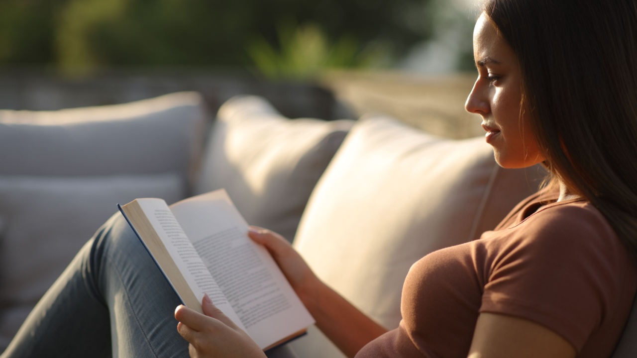 Woman reading a paper book sitting in a couch in a terrace at home