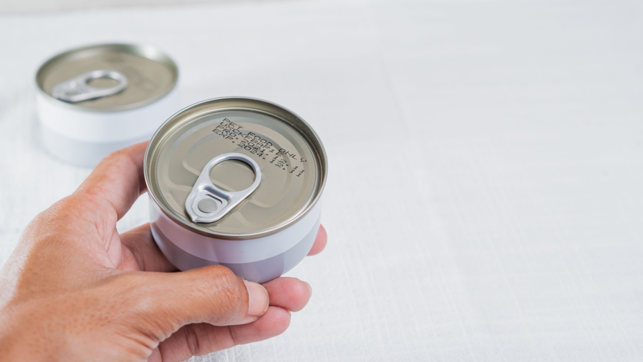 A person holding a can with a pull-tab lid, showing the bottom of the can. The background is a light-colored fabric, and there are additional cans slightly blurred in the