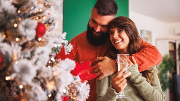 Beautiful young couple in love standing by nicely decorated Christmas tree, hugging and drinking champagne while relaxing at home on Christmas day