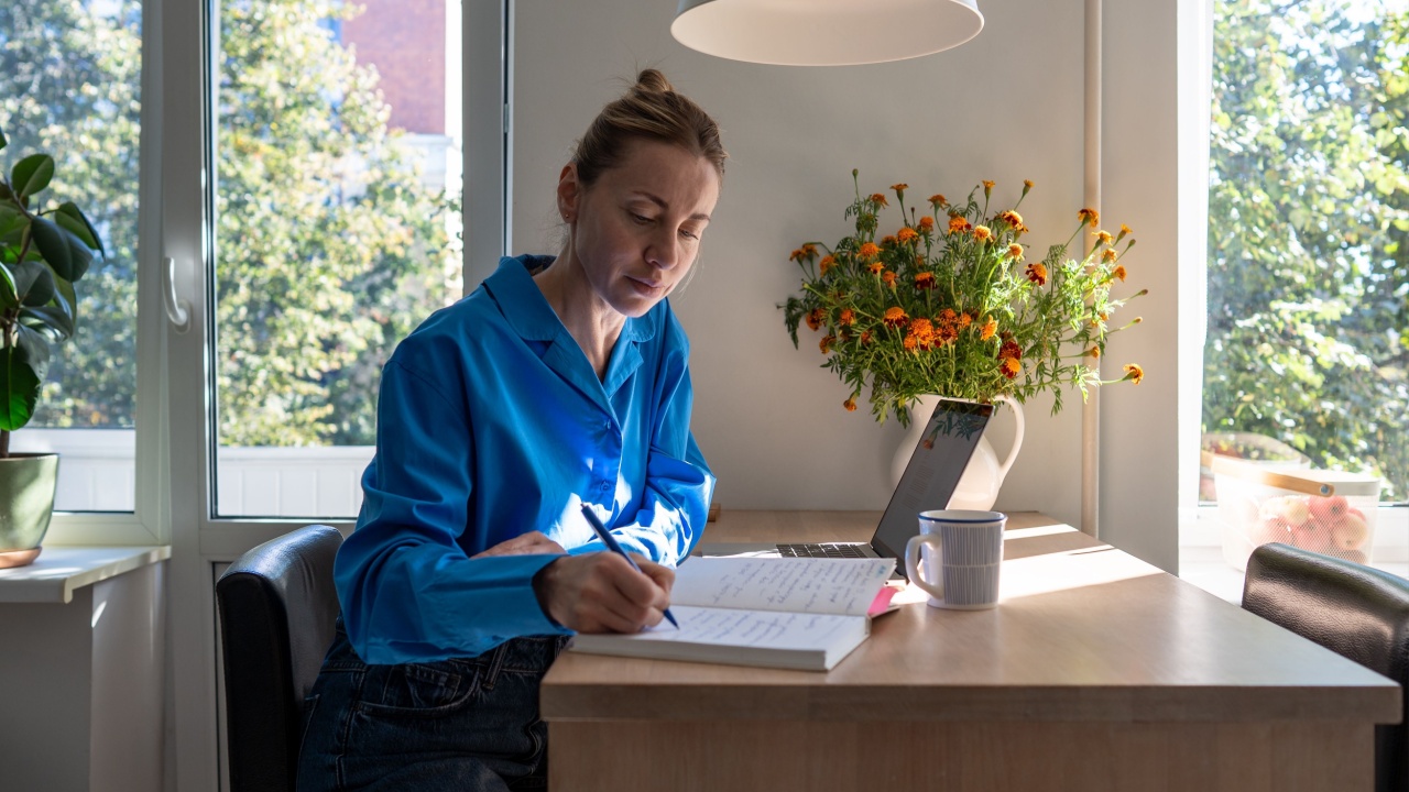 Concentrated middle aged woman works at home, planning day jotting down diary, comfortable sitting at table with laptop surround by houseplants. Focused female writing notes in day planner in morning