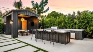 Modern outdoor kitchen and dining area with sleek black furniture and a stylish pergola, set against a lush green garden at sunset.