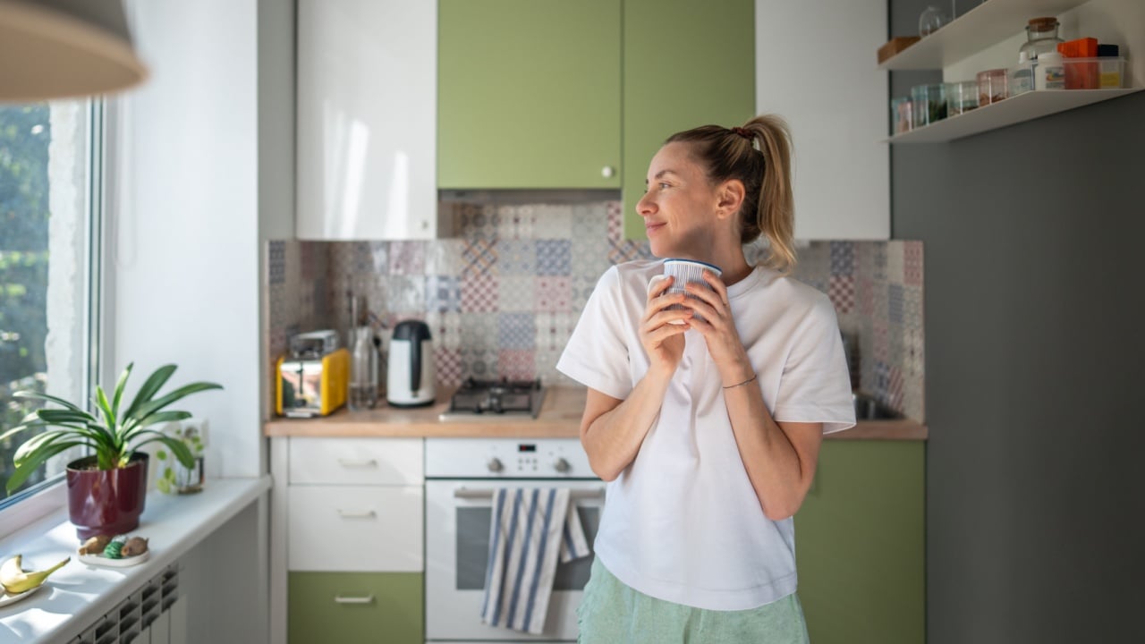 Joyful smiling woman with morning coffee, observing outside from domestic kitchen. Slow morning, contentment, relaxed mood, no rush weekend, mindful slow living, refreshing break, cozy routine.
