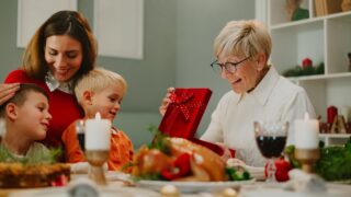 Happy family sitting at the dinner table at christmas time, enjoying their festive meal and celebrating while grandma opens a present