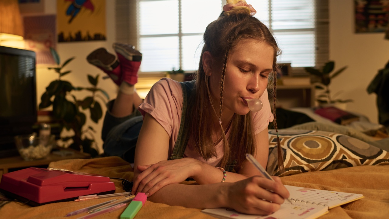 Young woman lying on bed and blowing bubble gum while writing in notebook during afternoon. Room decorated with plants, art, and fairy lights creating cozy retro atmosphere