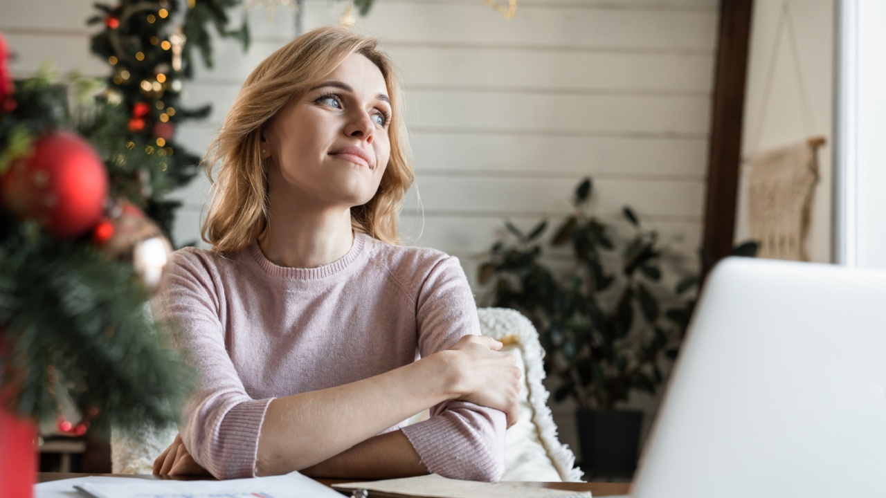 Young business woman at home with laptop on desk having a break sitting in living room with Christmas decoration tree thinking dreaming about winter holidays vacation looking aside