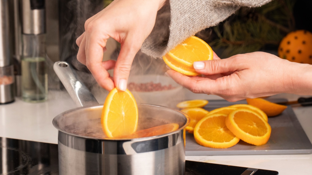 A woman adds a slice of orange to the boiling mulled wine in a saucepan in the kitchen.