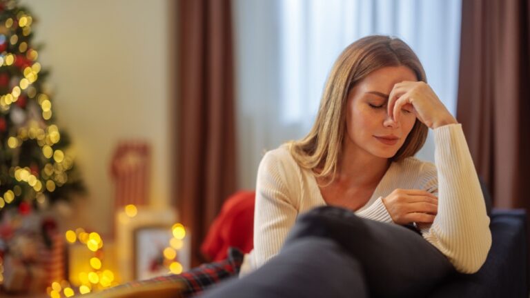 Young brunette woman is in her pajamas, sitting on a sofa, thinking about something during Christmas holiday and looking worried