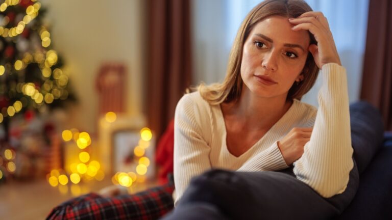 Young woman is in her pajamas, sitting on a sofa, thinking about something during Christmas holiday and looking worried