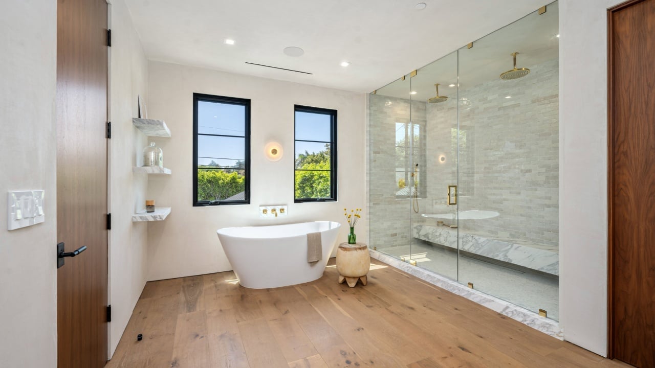Modern bathroom with freestanding tub, glass shower, and wooden floor, featuring natural light from large windows.