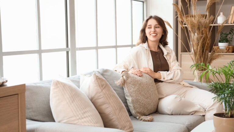Smiling housewife using handheld vacuum cleaner to remove dust and mites from sofa in living room, enjoying domestic chores and keeping her home clean and tidy