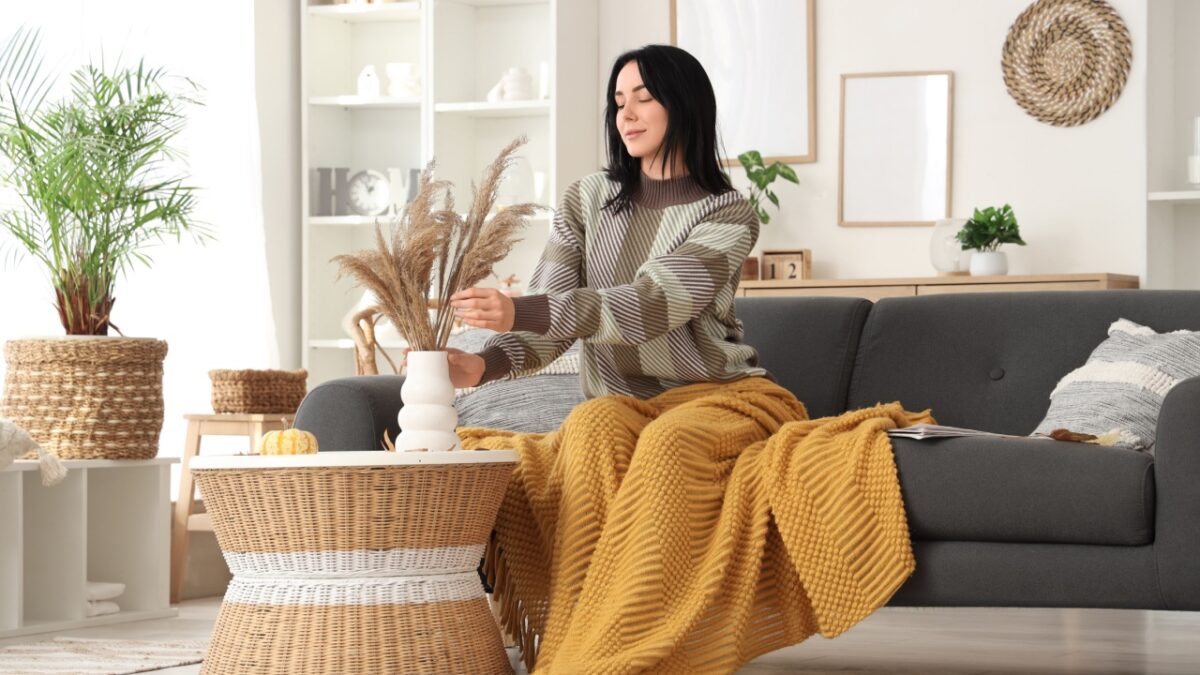 Beautiful young happy woman with warm plaid and vase sitting on sofa at home