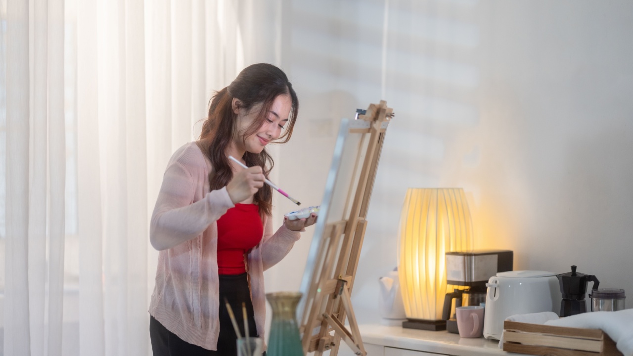 Young woman focuses intently on her artwork, painting on a canvas set up on an easel in her home studio. The scene captures a cozy, creative atmosphere with soft natural light filtering into the room