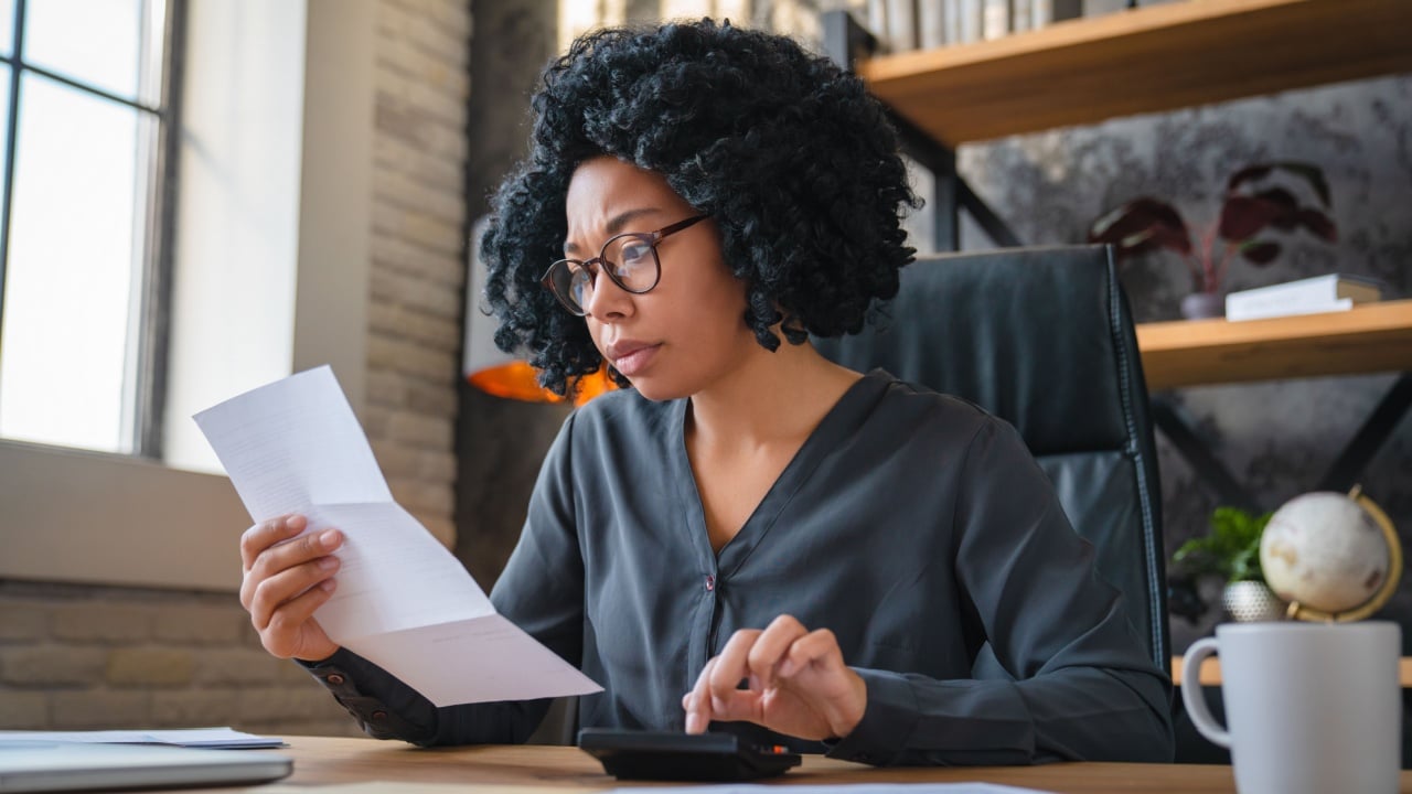 Focused millennial Black business woman calculating finance, money, using calculator, laptop computer at home workplace table, counting budget, paying bills, taxes, rent, mortgage fees