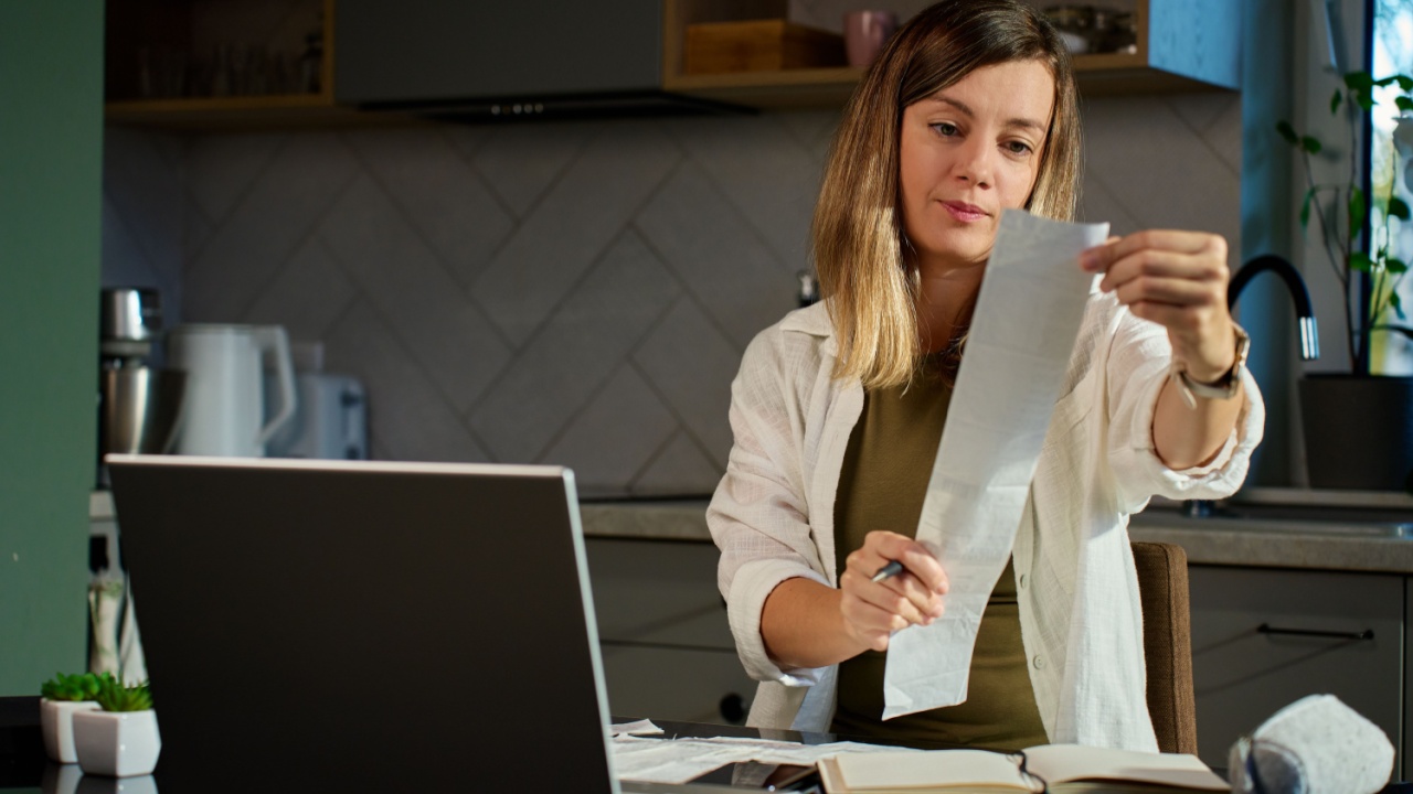 Stressed woman reviewing bills at home. Female organizing receipts, sitting at kitchen table with laptop and paper documents. Concept of budgeting and managing personal finances