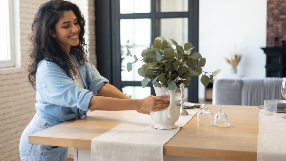 Modern home interior design, Scandinavian style concept. Beautiful young brunette woman arranging plants in vase, decorating dining room table, creating cozy atmosphere in her apartment