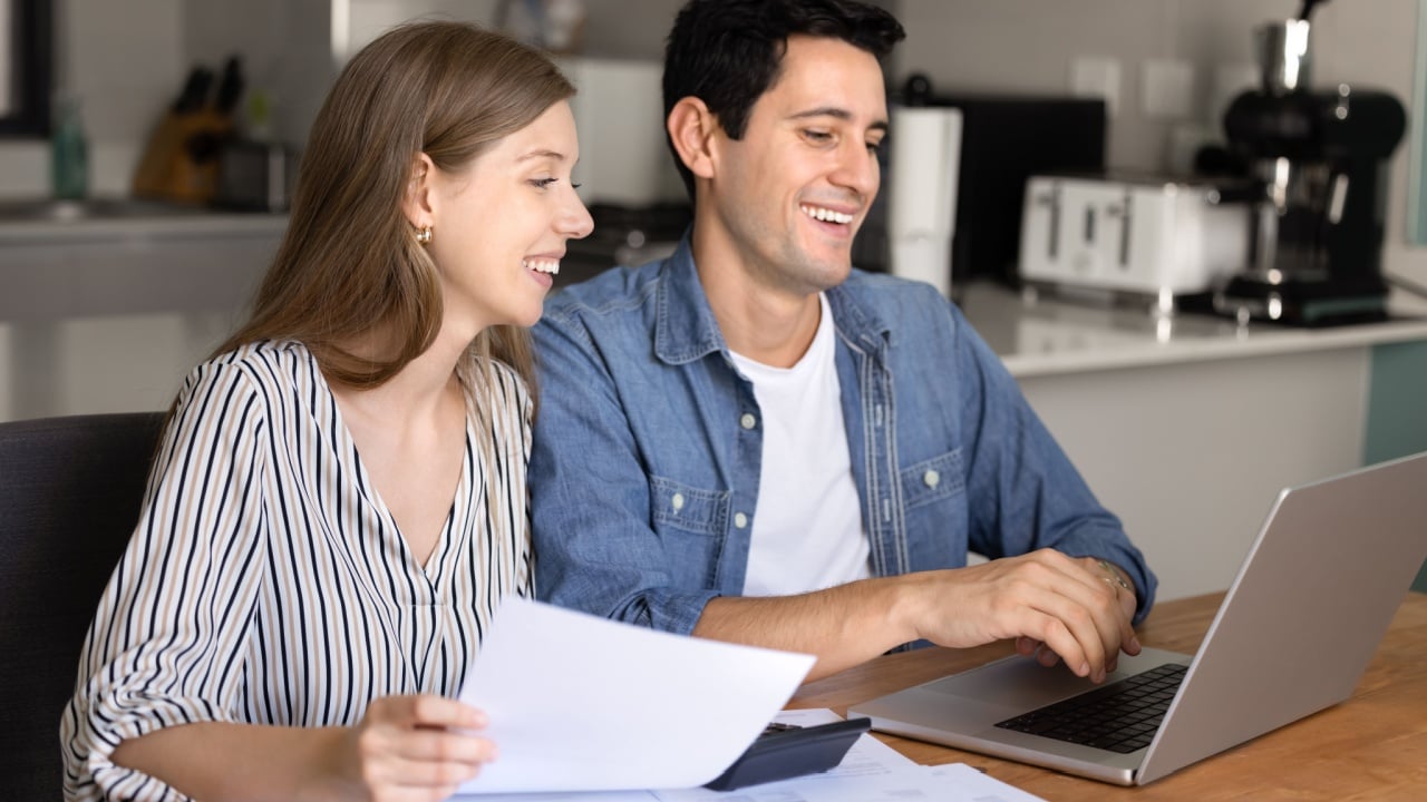 Happy financially successful young couple counting profit, checking income analytics, using bank application on laptop, checking invoices, paper bills for payment, smiling, laughing